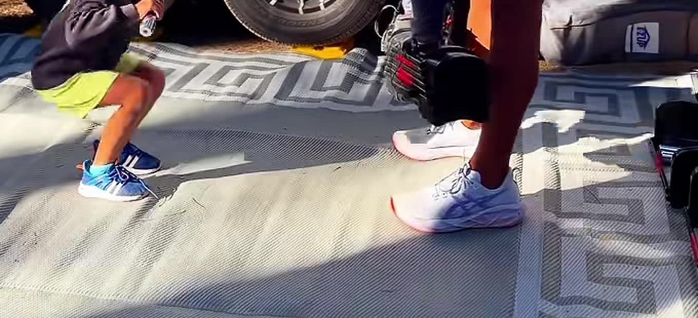 Kody's feet and dumbbell next to his young son's small sneakers, working out together on a rug outside an RV while camping.